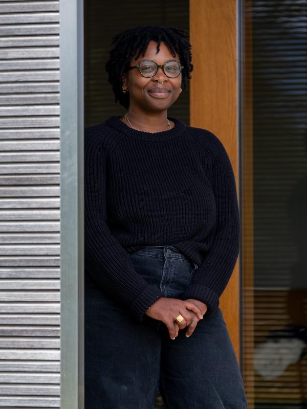 A photo of Zoé in a thick black sweater and black jeans, she has on a pair of round glasses; she's standing in front of a sliding glass door with a brown frame, her front shoulder is partially obscured by a metal column.